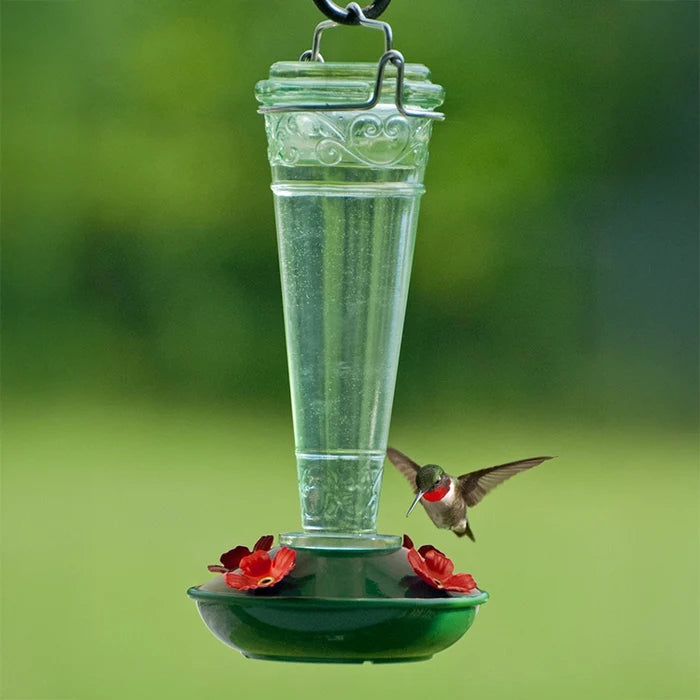 8 oz. Torchiere Glass Hummingbird Feeder with red flower ports and embossed green glass, attracting a hummingbird as it hovers to drink nectar.