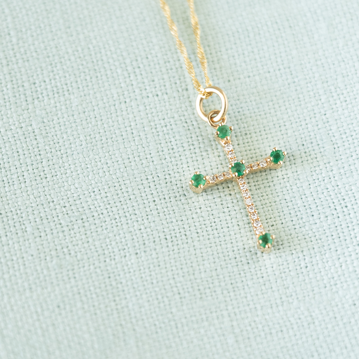 A gold cross pendant necklace with small green gemstones and clear stones, displayed on a textured light blue fabric background.