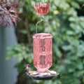 Rosewood Column Antique Hummingbird Feeder with ant moat hanging outdoors, featuring an embossed pink glass nectar reservoir and flower-shaped feeding ports.
